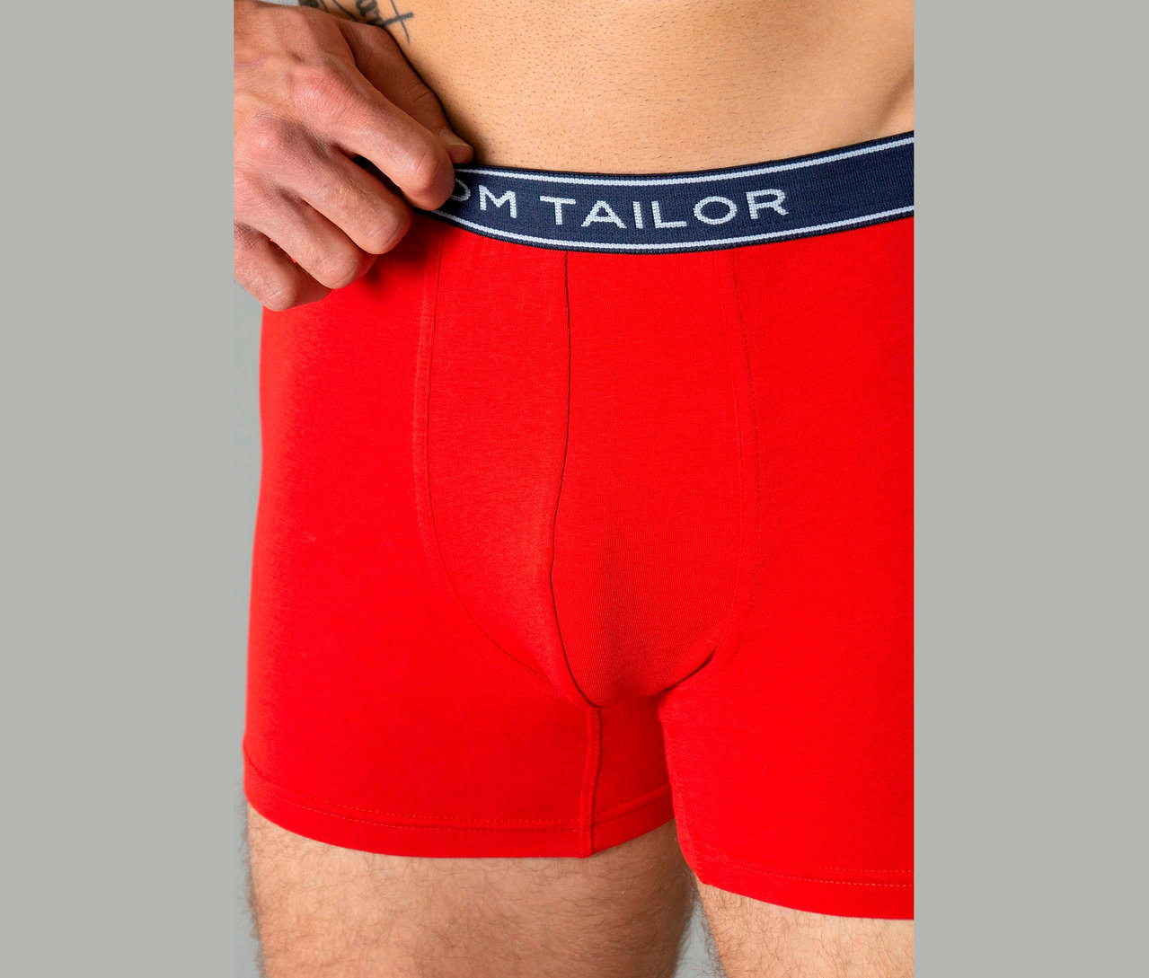 Muž má na sobě červené boxerky s tmavě modrým pasem s textem „TOM TAILOR“.