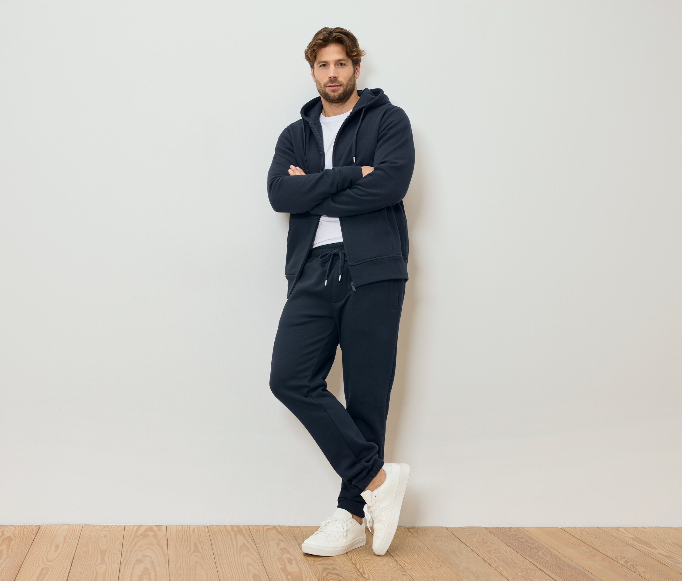 Muž se zkříženýma rukama má na sobě tmavě modrou mikinu s kapucí na zip, tmavě modré tepláky a bílé tenisky, stojí před bílou stěnou.