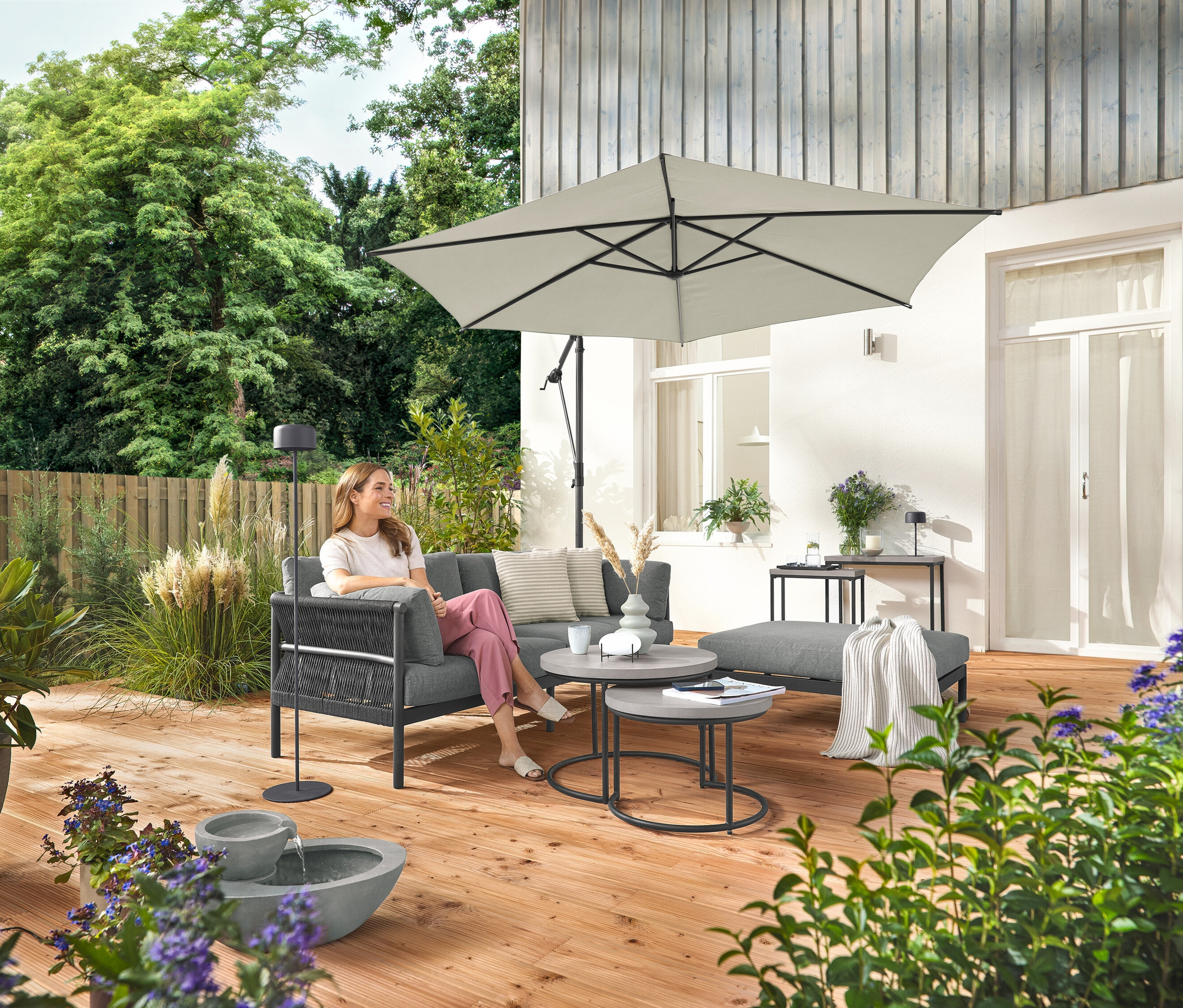 Žena sedí na pohodlné zahradní pohovce se samostatnou podnožkou. Vedle ní stojí šedá solární dobíjecí stojací lampa. V popředí je solární zahradní fontána.