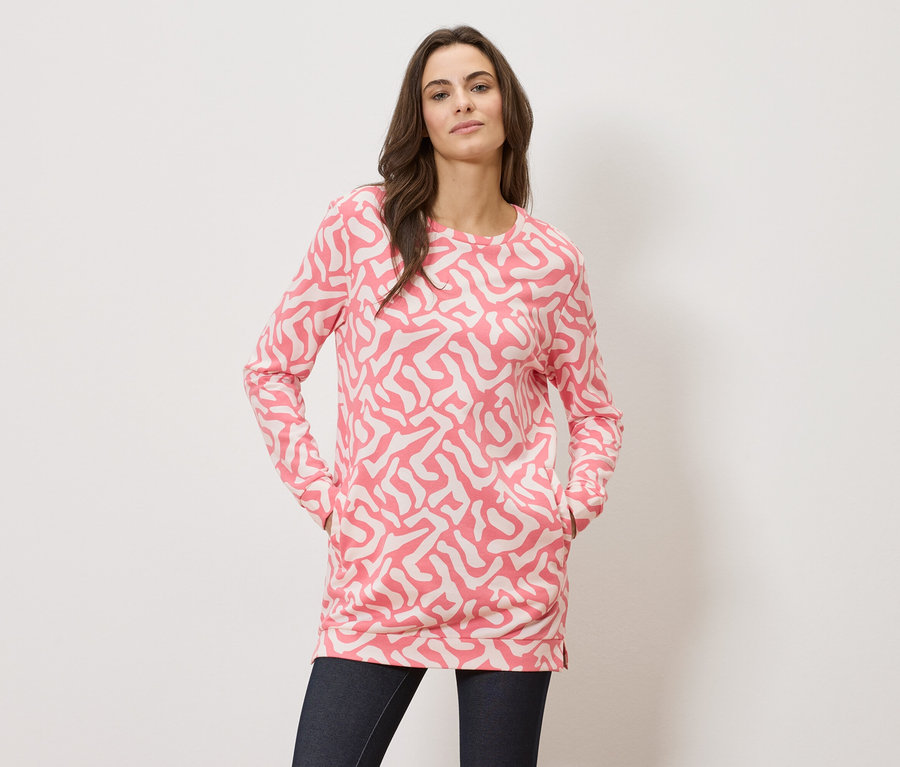 Žena s rukama v kapsách má na sobě top s dlouhým rukávem s růžovým vzorem a tmavé džíny.