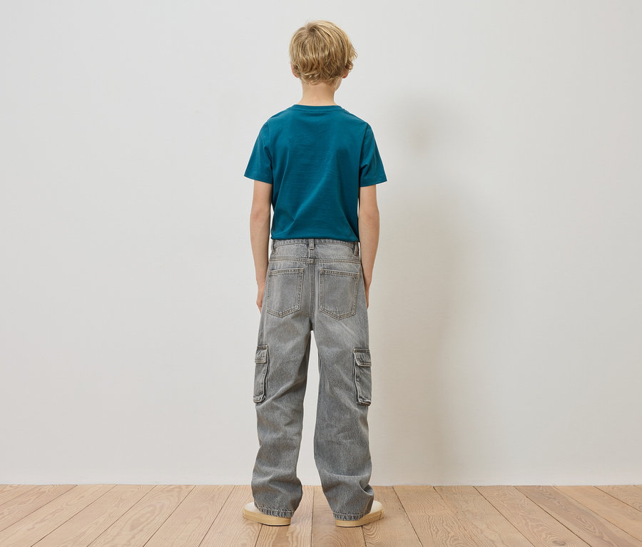 Chlapec s blond vlasy stojí zády ke kameře, má na sobě modrozelené tričko a šedé cargo džíny.
