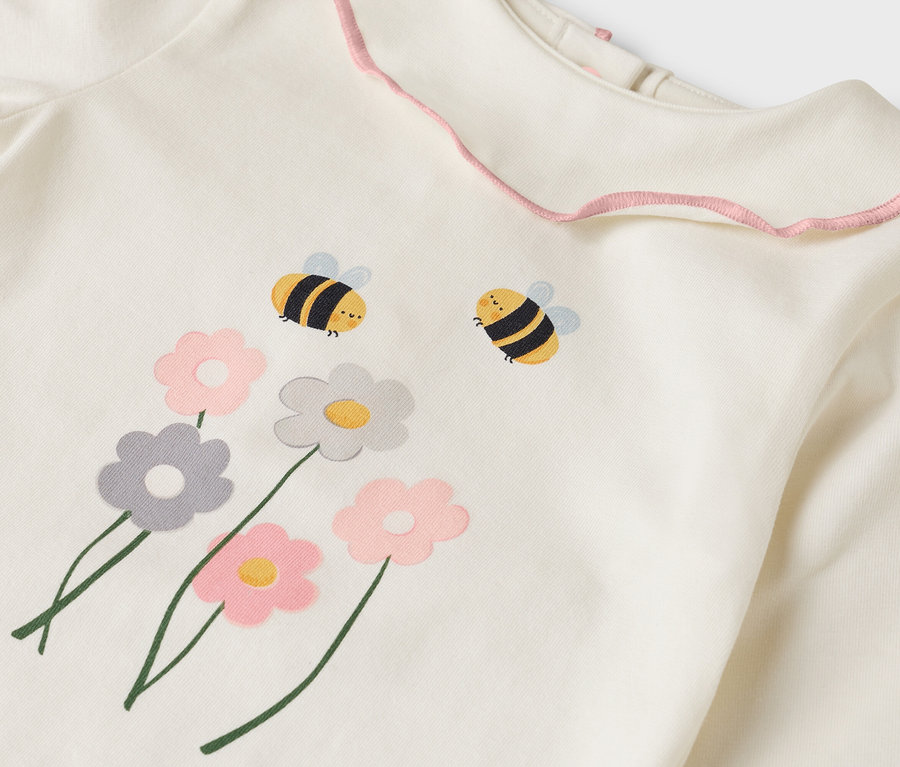 Detailní pohled na krémový top s růžovým volánkovým límcem, zdobený květinovými a včelími motivy.