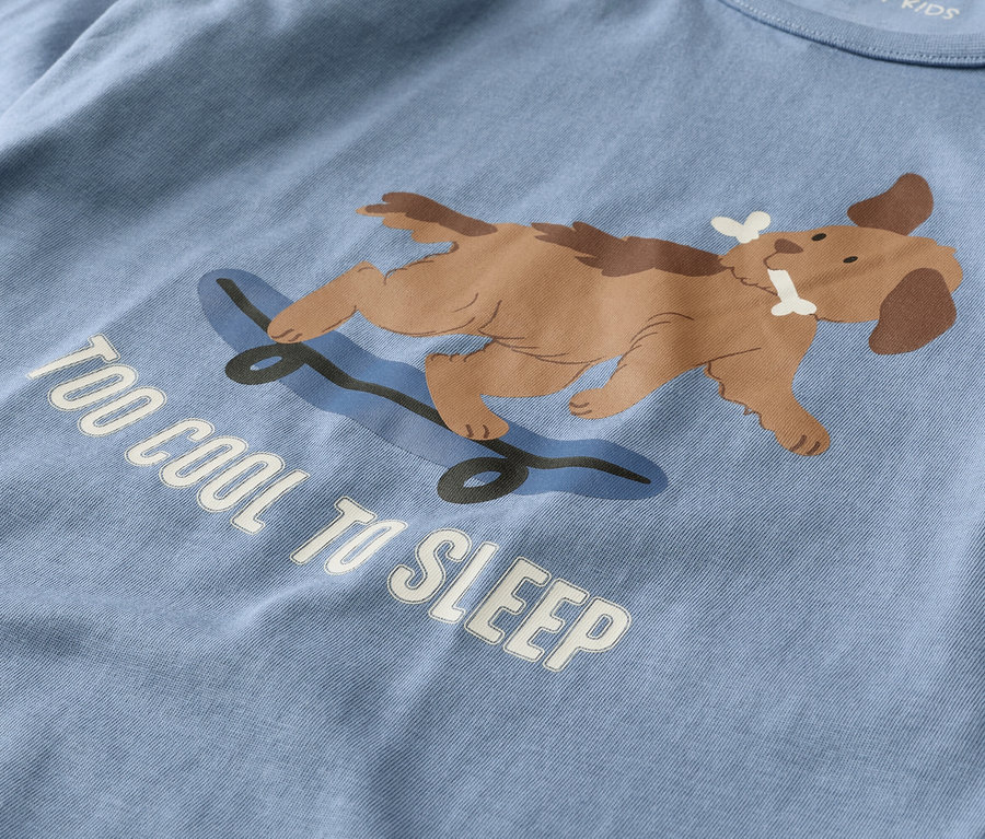 Světle modré tričko s hnědým psem ležícím na modrém skateboardu a držícím kost v tlamě. Pod ním je bílý nápis "TOO COOL TO SLEEP".