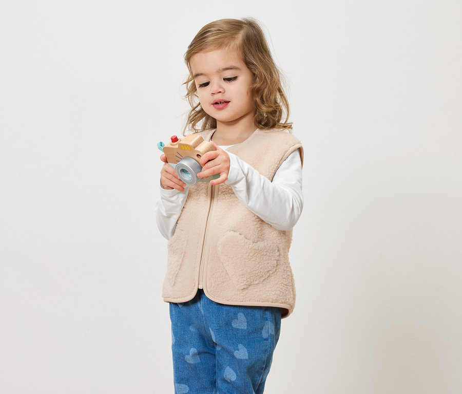 Malá holčička s kudrnatými vlasy drží hračkový fotoaparát na světlém pozadí.