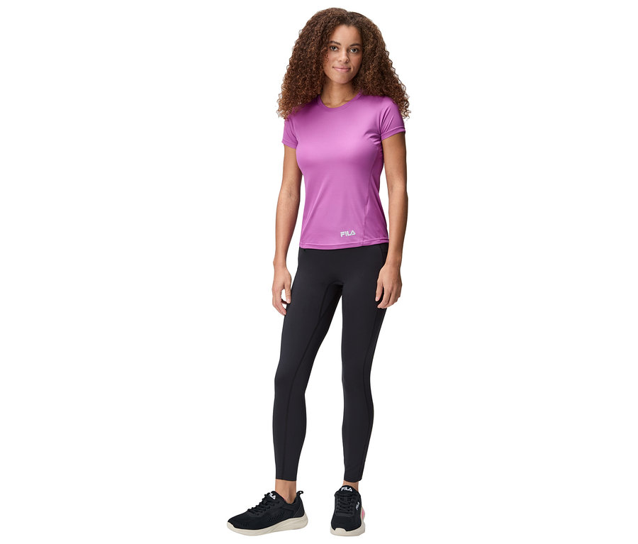 Žena stojí a má na sobě fialové tričko Fila, černé legíny a černé tenisky.