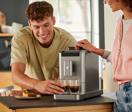 Muž sleduje, jak se káva lije do dvou sklenic z plnoautomatického kávovaru »Esperto2 Caffè« v titanově stříbrné barvě.