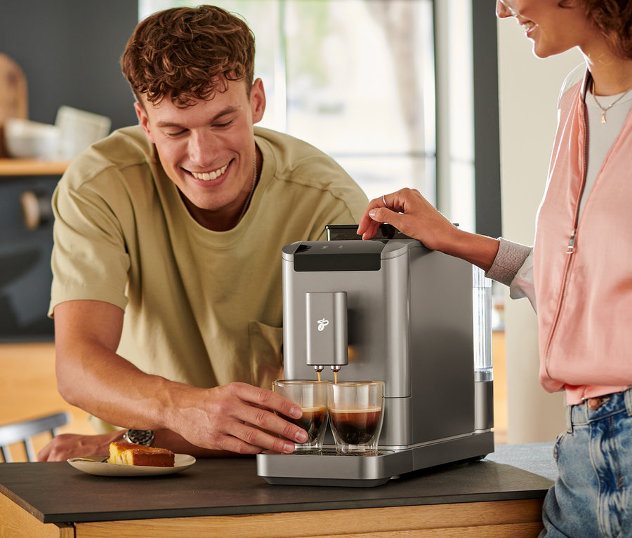 Muž sleduje, jak se káva lije do dvou sklenic z plnoautomatického kávovaru »Esperto2 Caffè« v titanově stříbrné barvě.