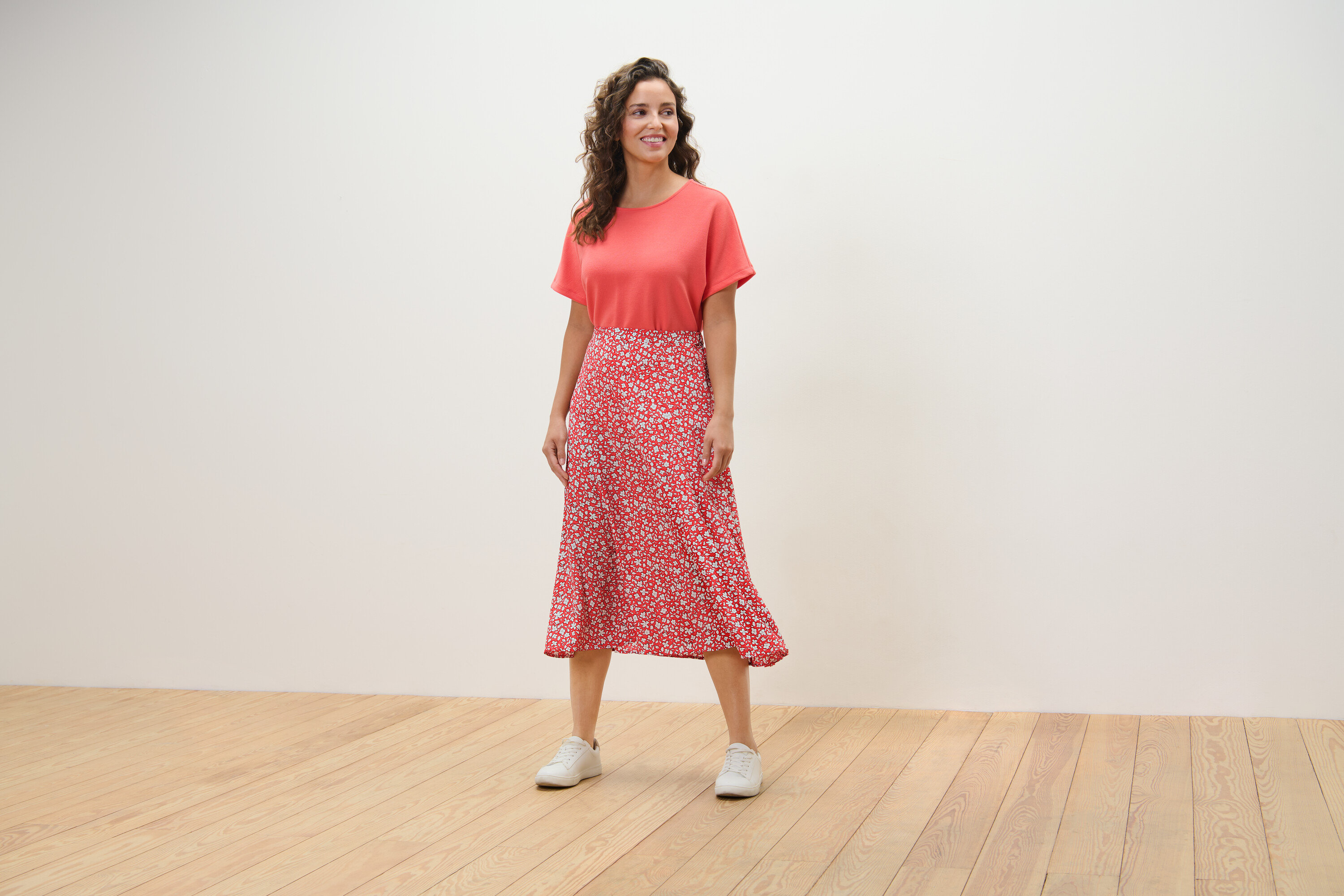 Žena stojící na dřevěné podlaze má na sobě korálovou sukni s potiskem.