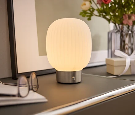 Noční lampa s bílým stínidlem stojí na skříňce vedle brýlí, knihy, vázy s květinami a dárkové krabičky.