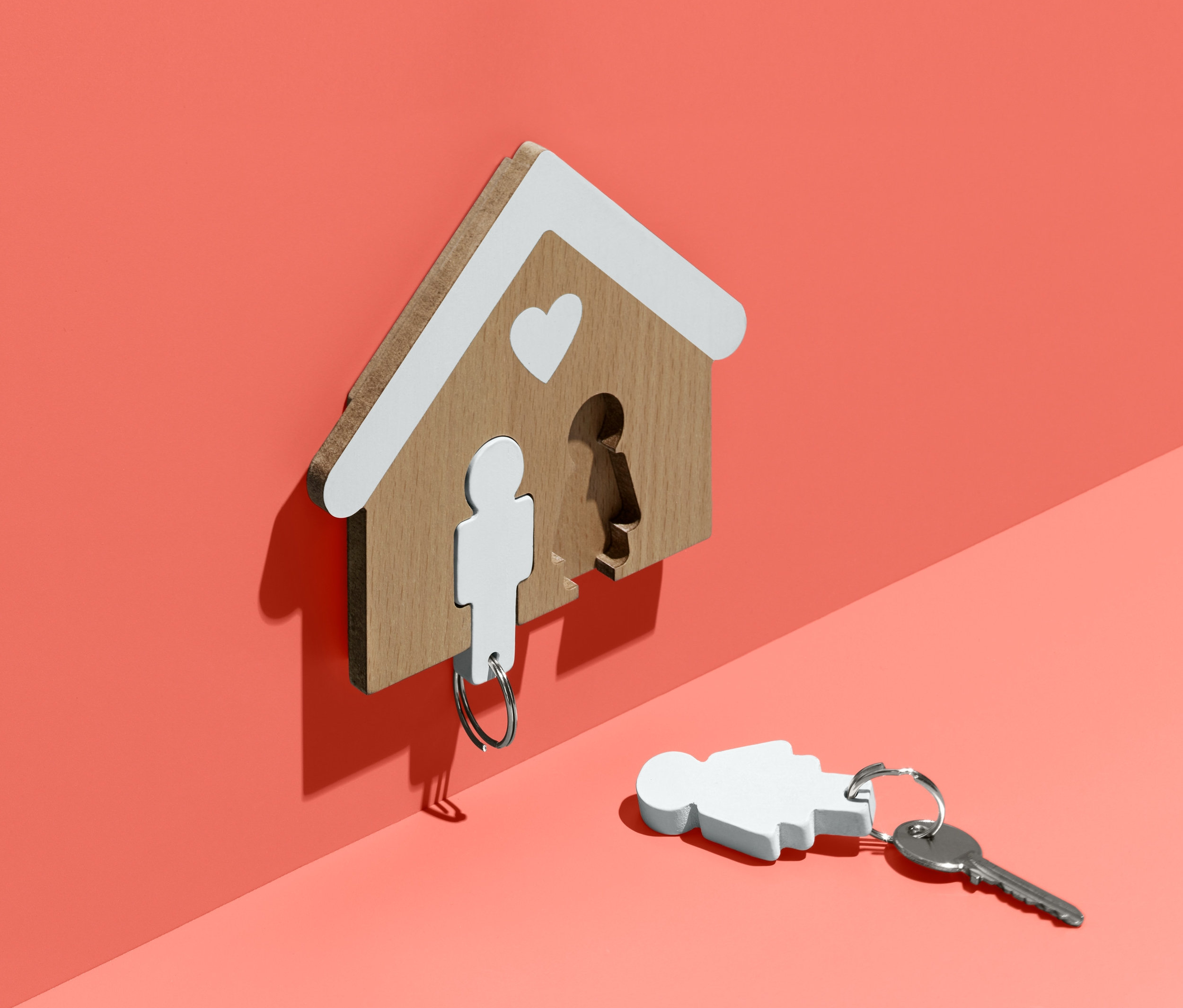 Na korálové zdi visí dřevěný věšák na klíče ve tvaru domu s bílou střechou a srdcem. Jeden klíč s přívěskem ve tvaru postavy visí z věšáku, druhý leží na zemi.