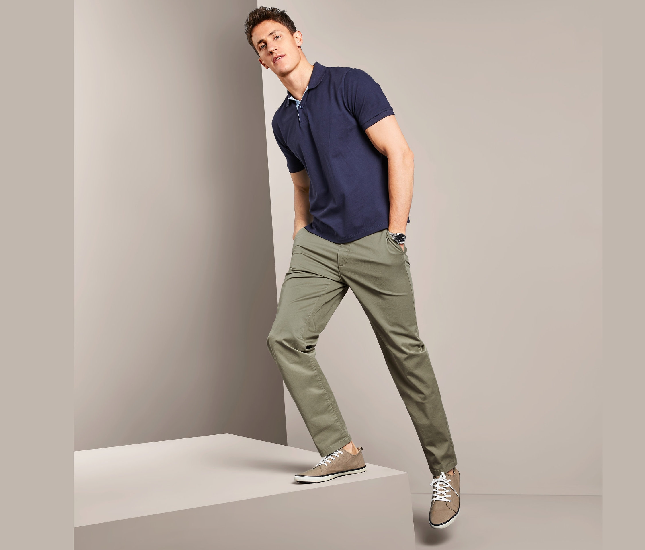 Muž v tmavě modrém tričku a khaki kalhotách chino stojí na šedém pódiu.