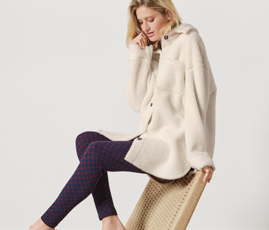 Žena sedí na židli a má na sobě dlouhé triko z hebkého plyše krémové barvy a vzorované legíny.