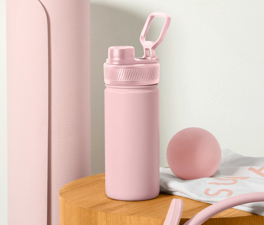 Růžová sportovní láhev na pití stojí na dřevěném stolku vedle srolované podložky na cvičení.