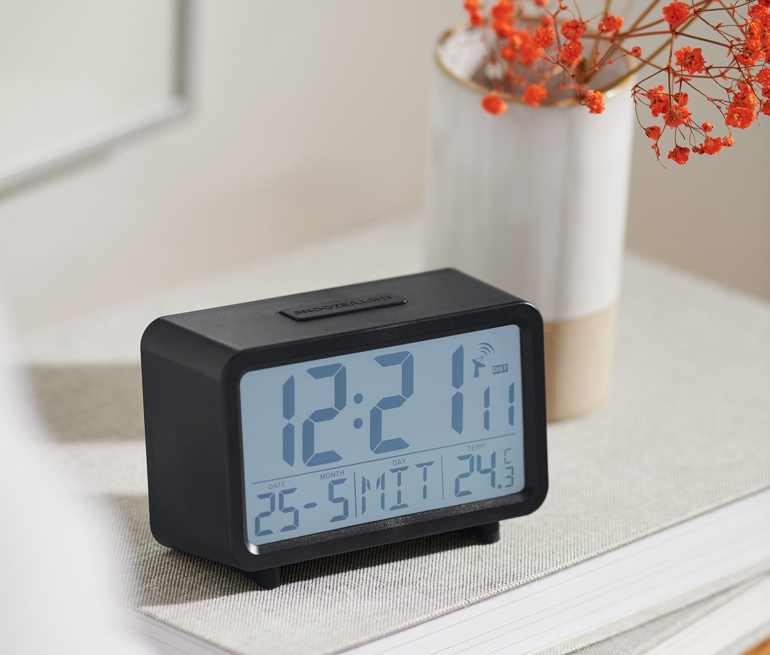 Černý digitální LCD budík řízený rádiovým signálem stojí na knize vedle vázy s oranžovými květy.