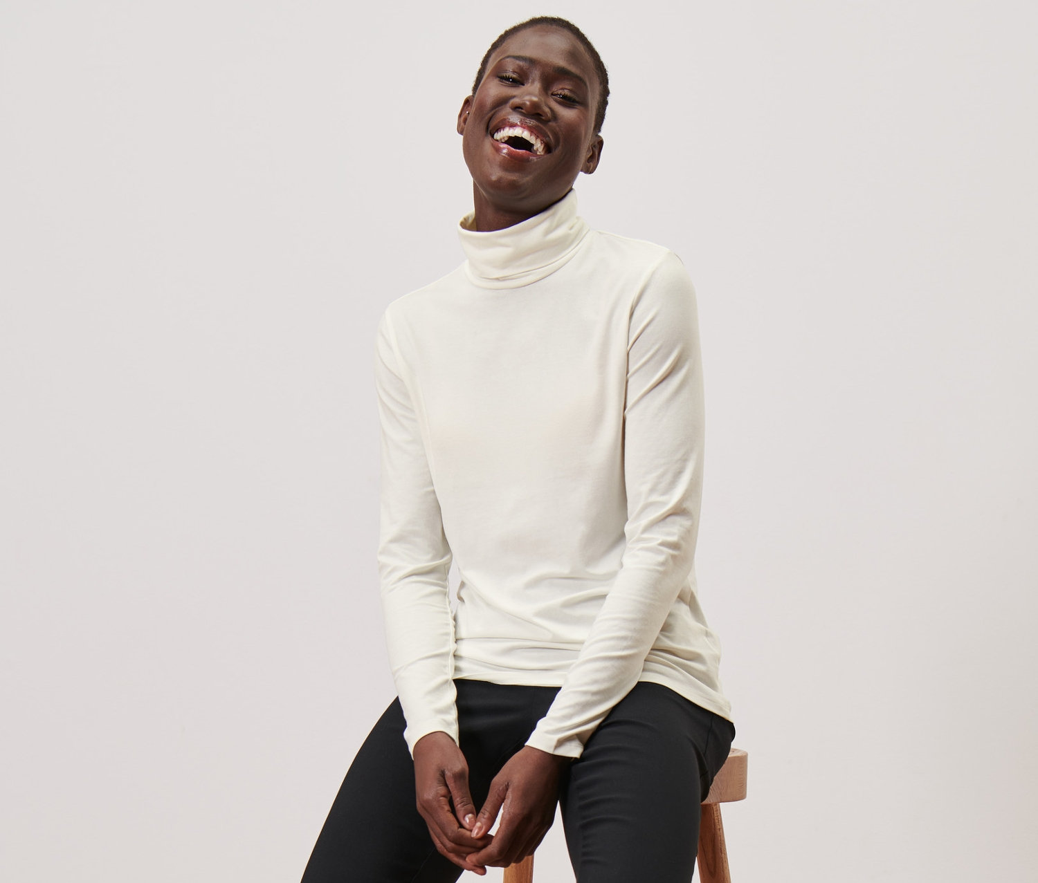 Žena sedí na stoličce a má na sobě krémové triko s rolákem, černé kalhoty a černé mokasíny.