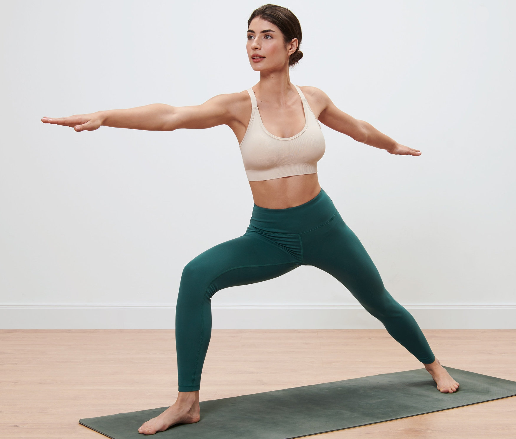 Žena v legínách a sportovní podprsence stojí na podložce na jógu a má rozpažené ruce.