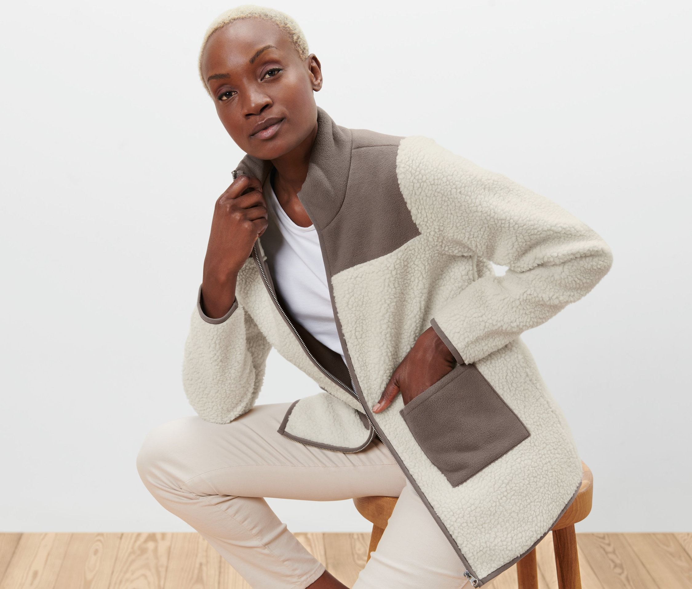 Žena sedí na stoličce a má na sobě béžovo-hnědý plyšový kabátek.