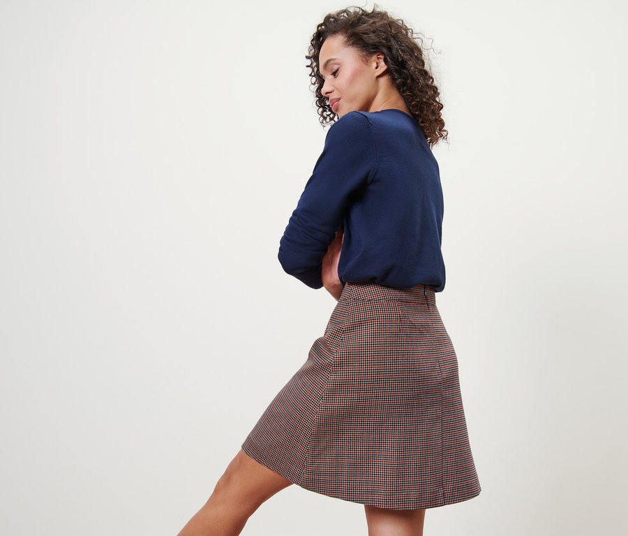 Žena pózuje v kostkované tkané sukni a modrém svetru.