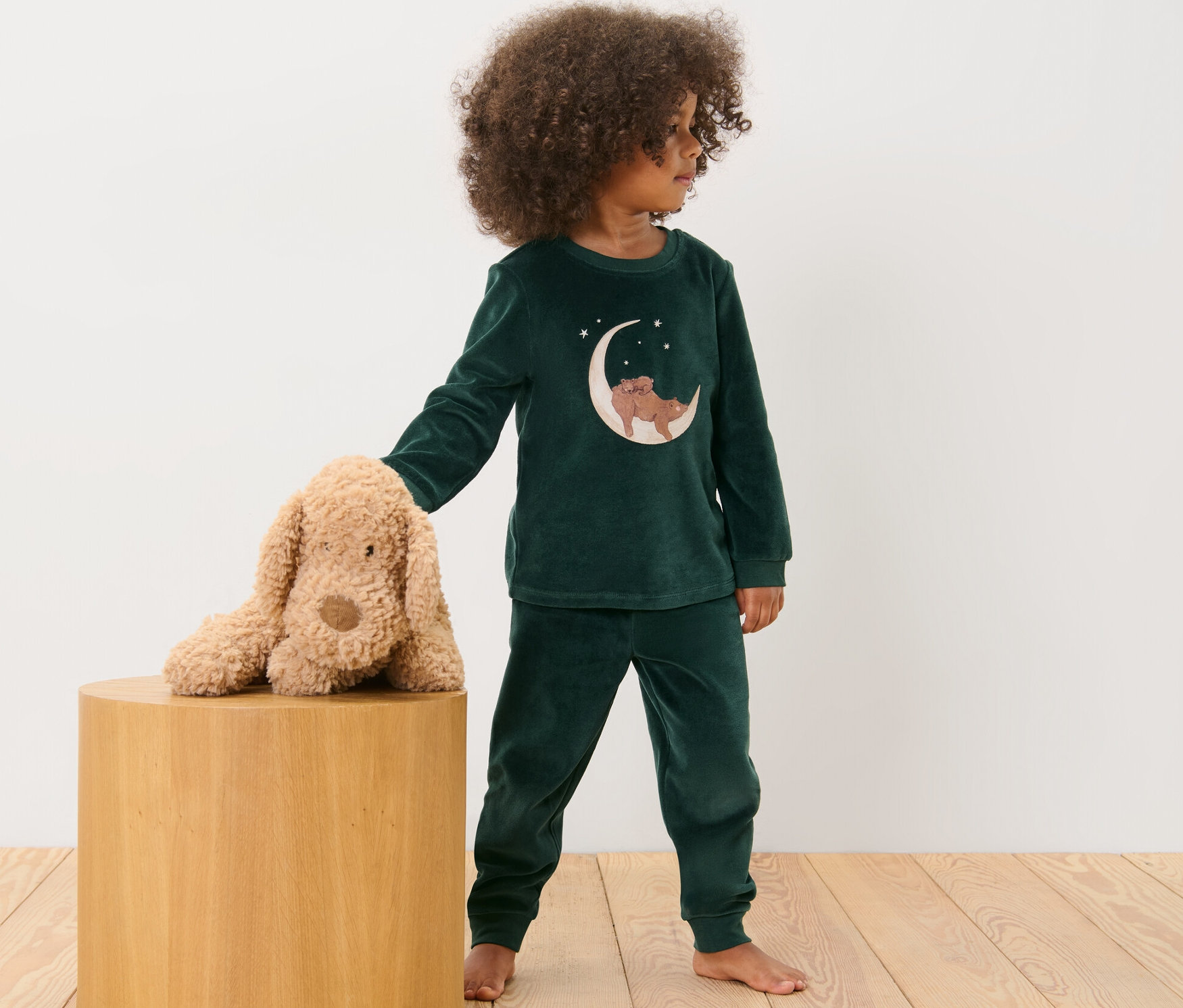 Dítě v plyšovém pyžamu stojí vedle plyšového psa na dřevěné stoličce.