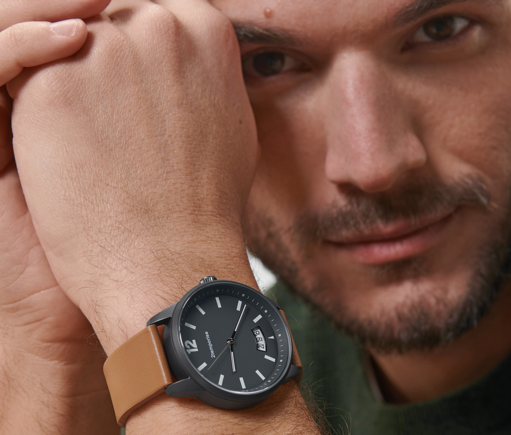 Muž se dívá do kamery a na zápěstí má černé náramkové hodinky v leteckém stylu s hnědým koženým páskem.