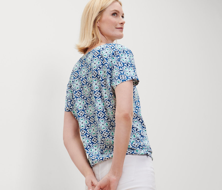 Žena s blond vlasy pózuje v halence s celoplošným potiskem a bílých kalhotách.