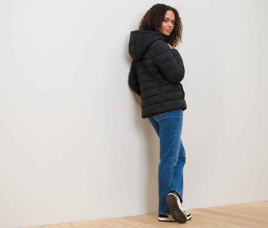 Žena se opírá o zeď a má na sobě prošívanou bundu s kapucí.