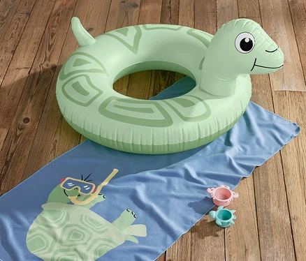 Zelený plavecký kruh Želva leží na modré osušce s potiskem želvy a dvou hračkách na dřevěné podlaze.