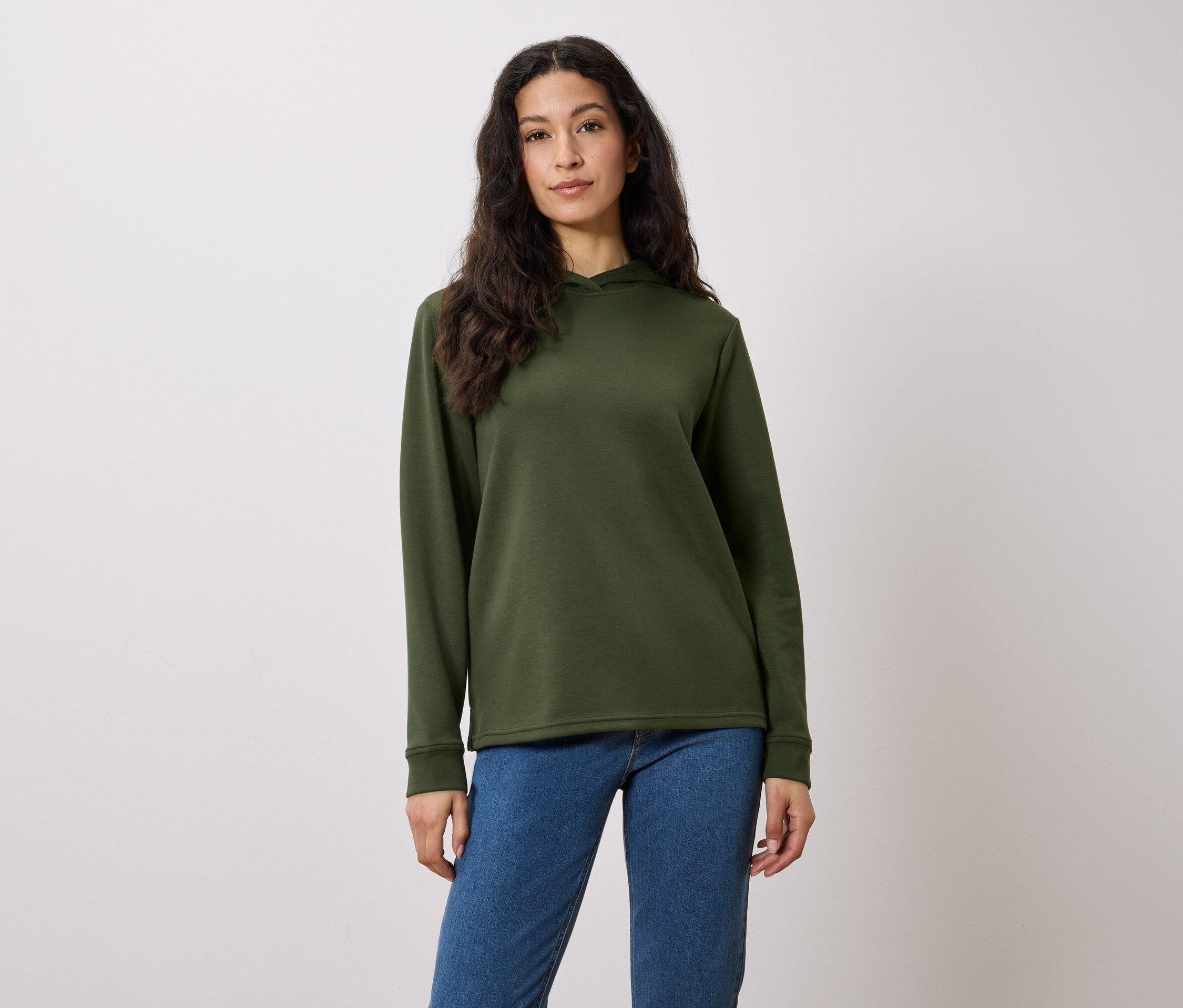 Žena s dlouhými hnědými vlasy pózuje v olivově zelené mikině s kapucí a modrých džínách před bílým pozadím.