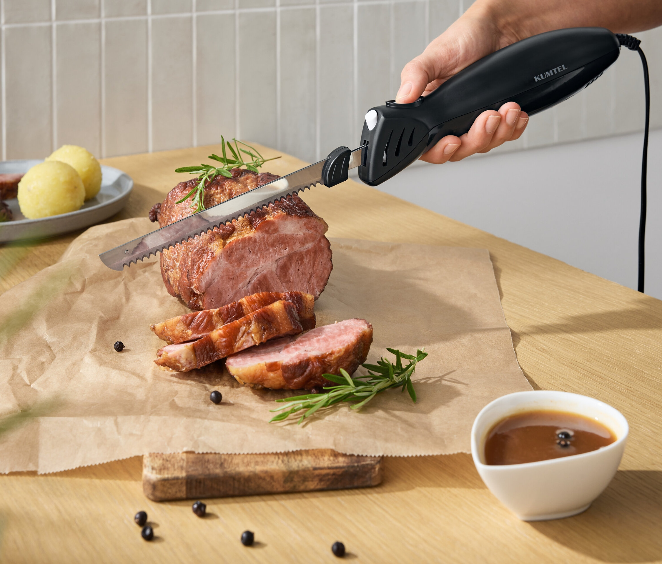 Ruka krájí elektrickým nožem pečené maso na prkénku. Na stole je omáčka a příloha.