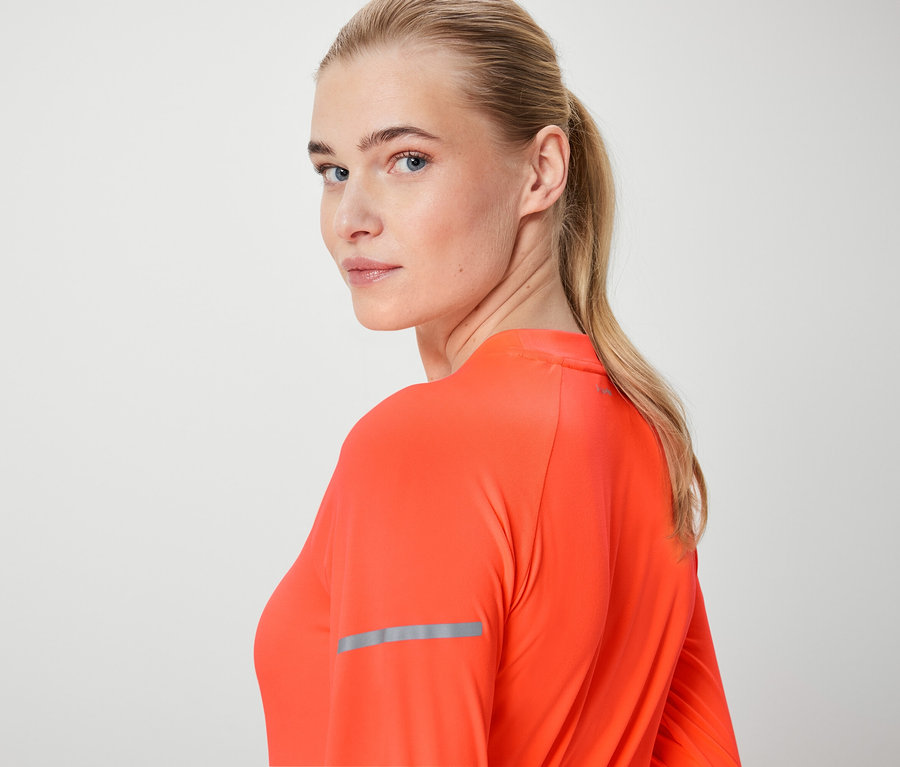 Blonďatá žena se dívá přes rameno, má na sobě oranžový top s dlouhým rukávem.