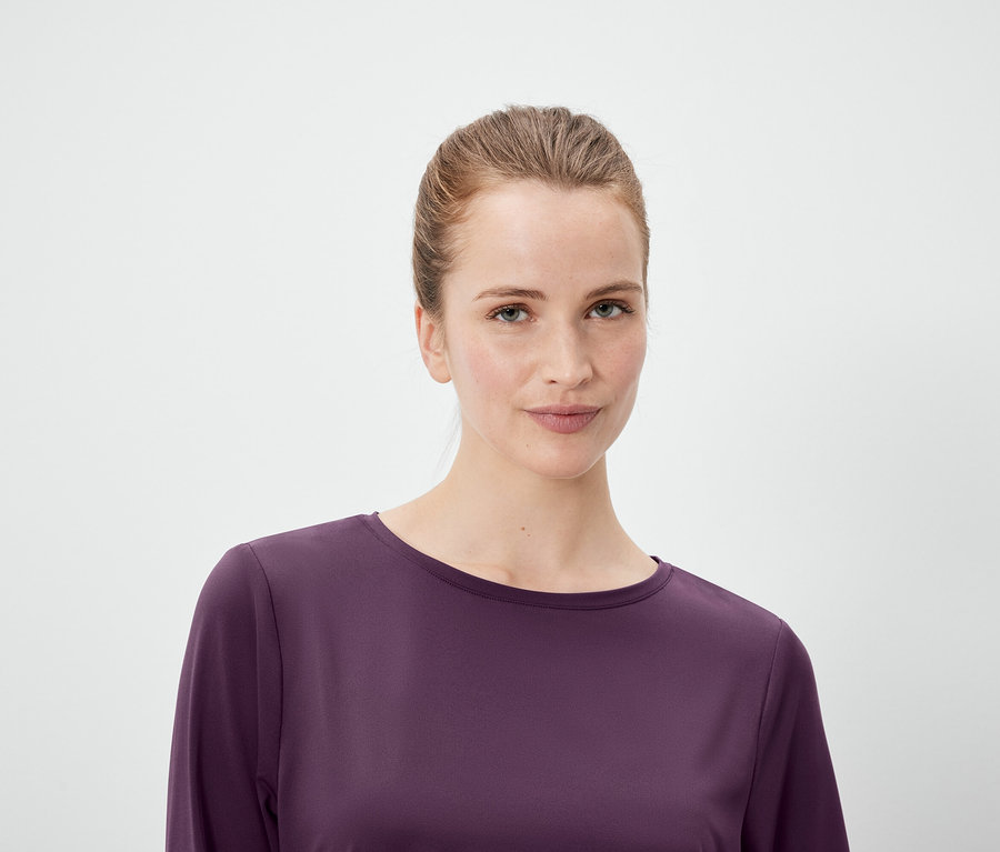 Portrét ženy s vlasy staženými do culíku, oblečené do fialového topu s dlouhým rukávem, na bílém pozadí.