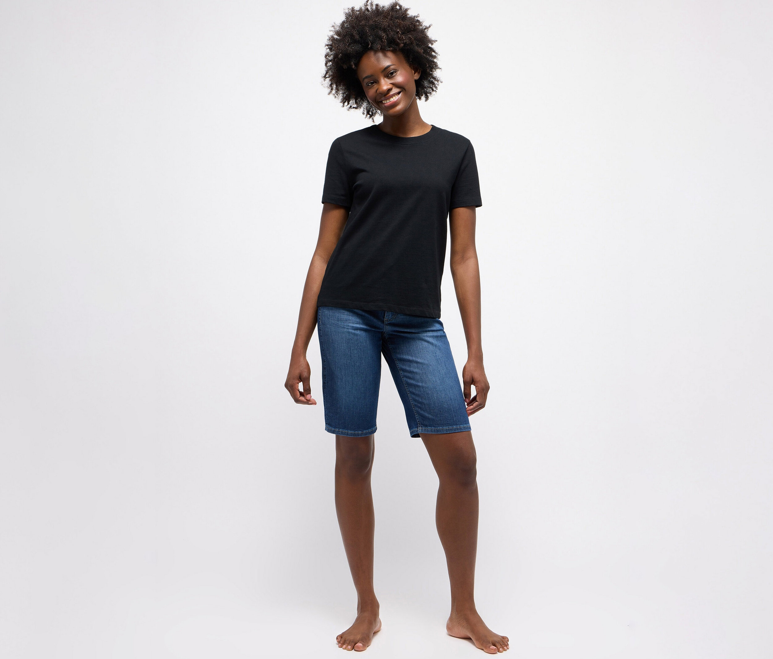 Žena stojí bosá před bílým pozadím a má na sobě černé tričko a džíny Angels »Bermuda Turn-Up«, straight fit, mid blue.