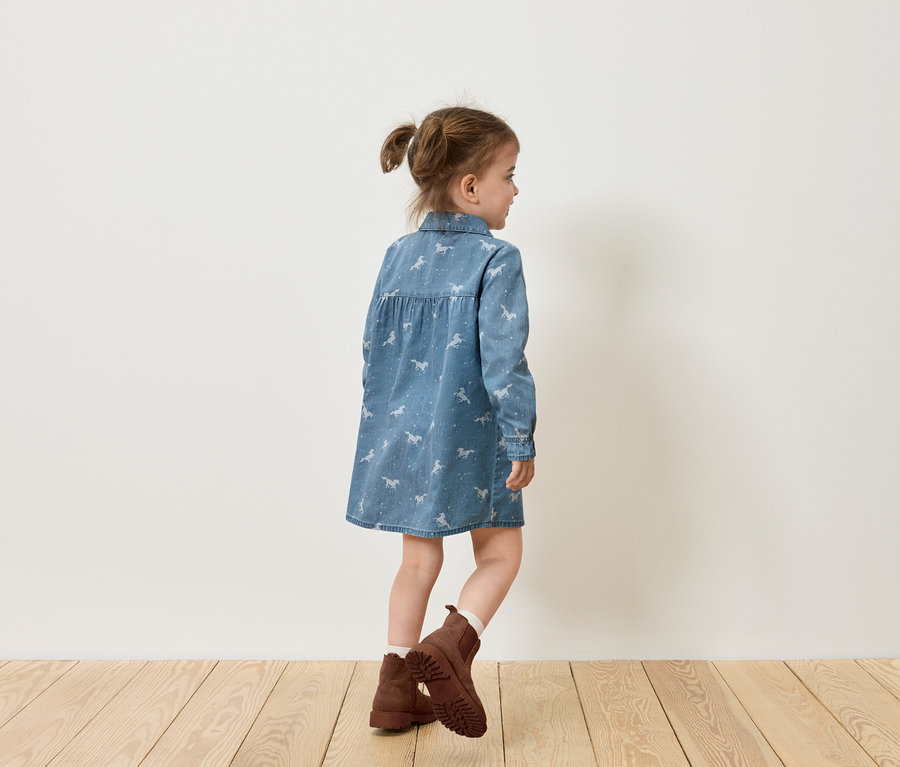 Dívka v džínových šatech s potiskem koní a hnědých botách stojí bokem k bílé stěně.