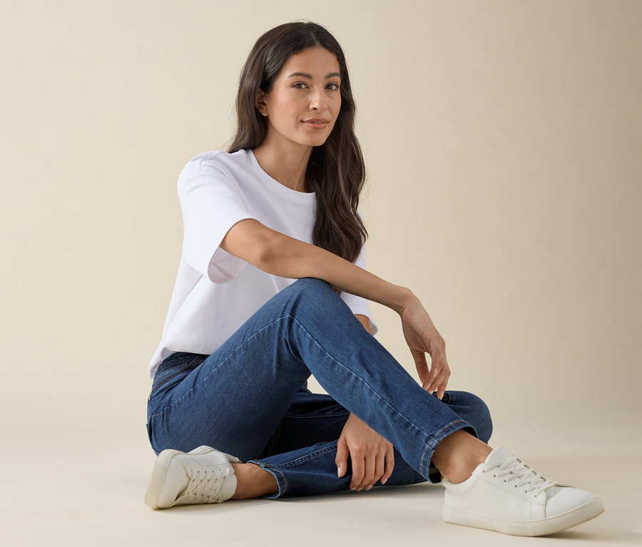 Žena sedí na zemi v džínách slim střihu »Emma« v barvě mid blue denim, bílém tričku a teniskách.
