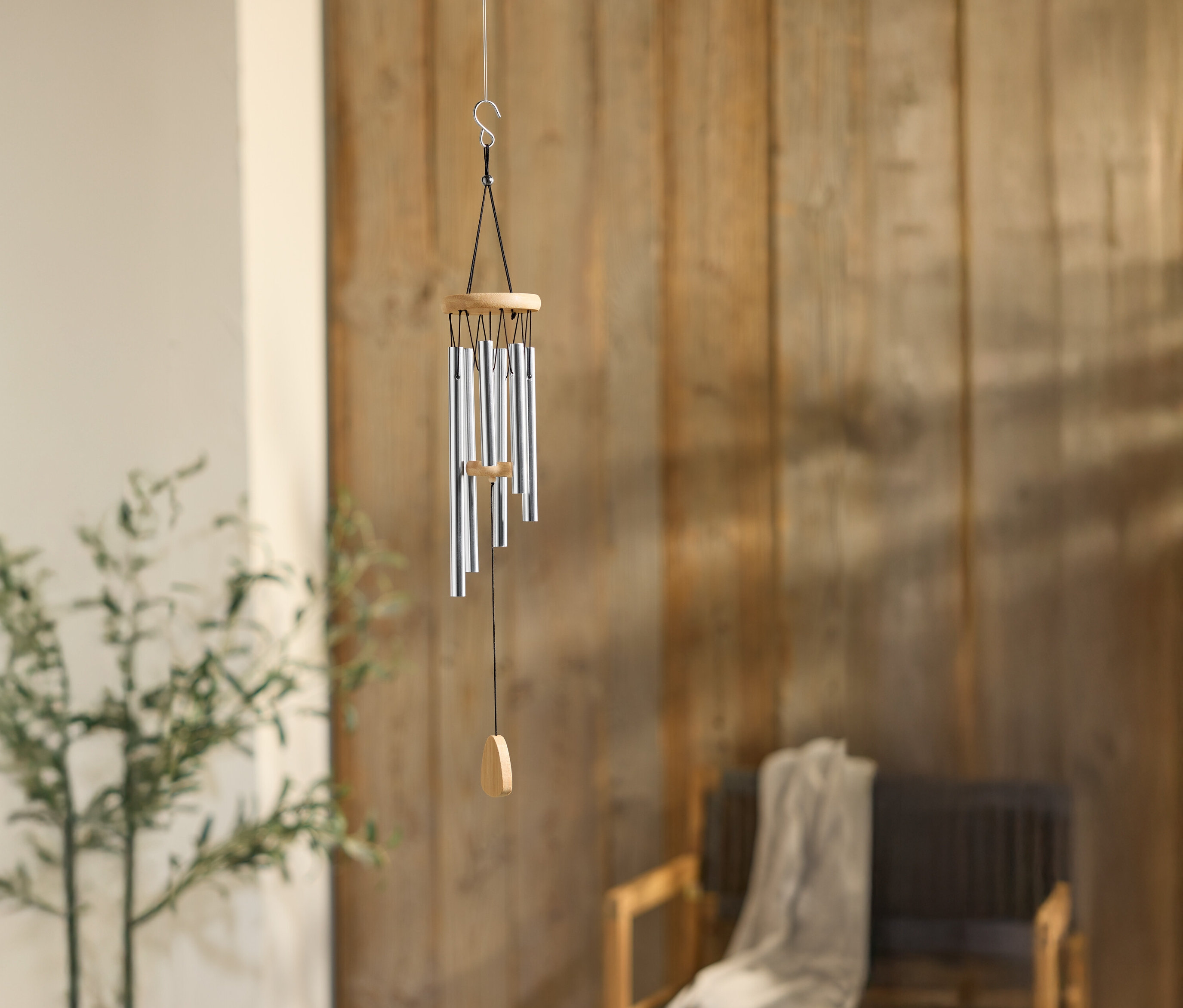 Větrná zvonkohra se stříbrnými trubkami visí před dřevěnou stěnou, vedle rostliny a židle.