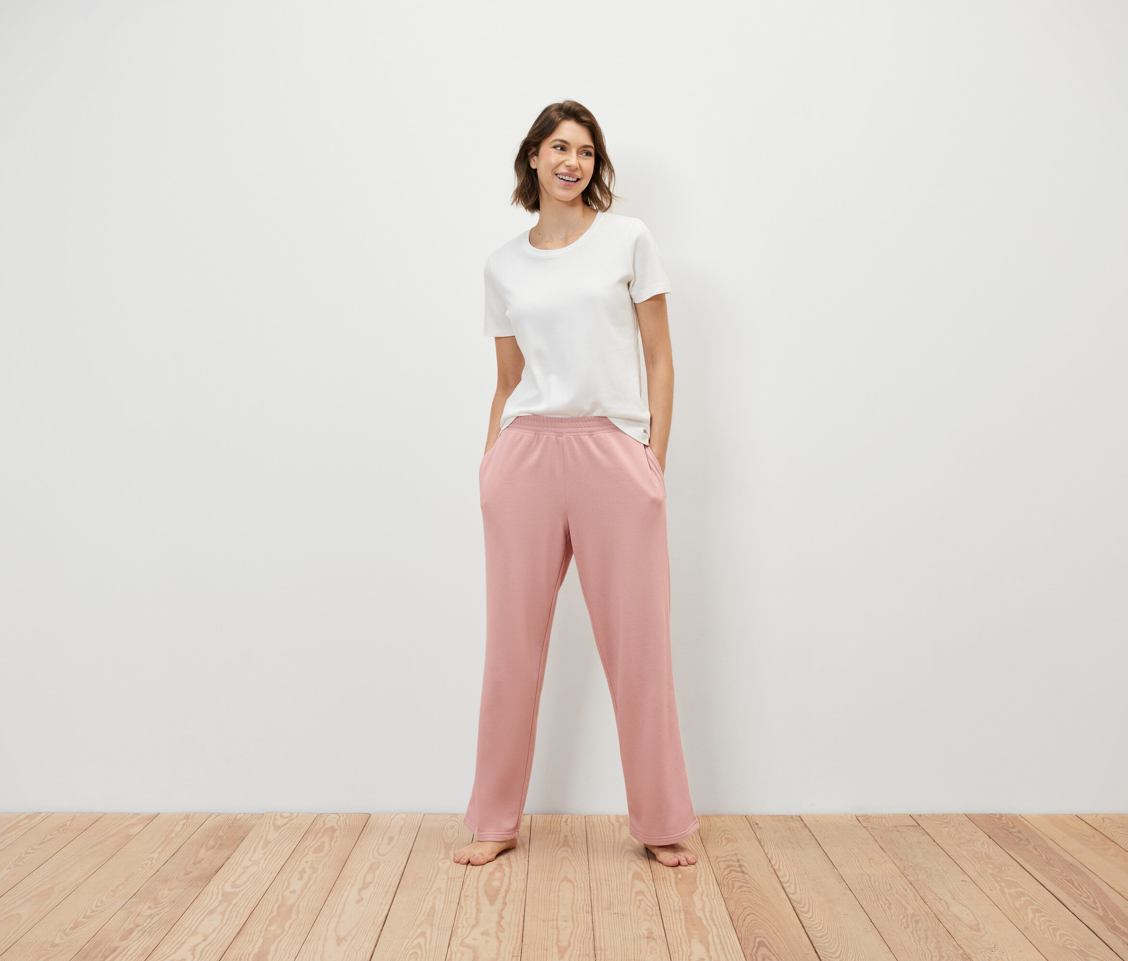 Žena stojí a má ruce v kapsách růžových tepláků a na sobě bílé tričko.