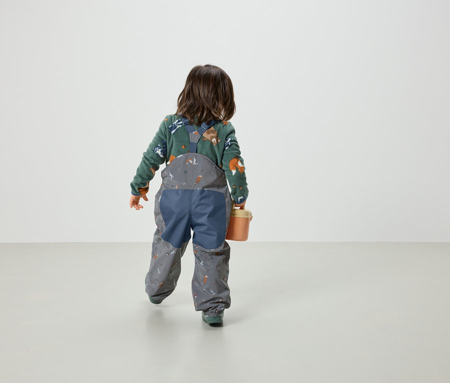 Dítě zády k fotoaparátu v zeleném tričku s potiskem lišek a šedých kalhotách s laclem a potiskem. Dítě drží oranžový kyblík.