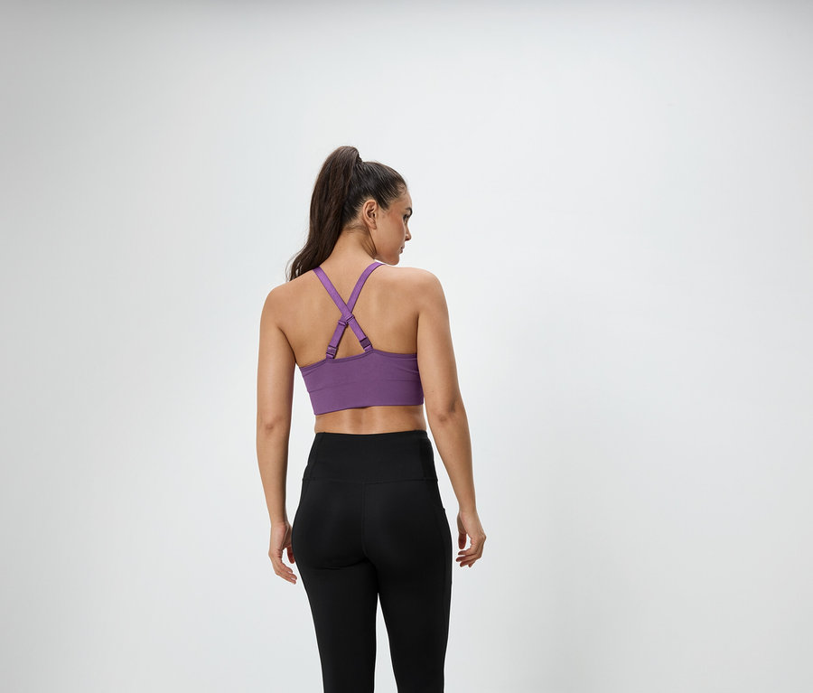 Žena s hnědými vlasy staženými do culíku stojí zády k fotoaparátu a má na sobě fialovou sportovní podprsenku a černé legíny.