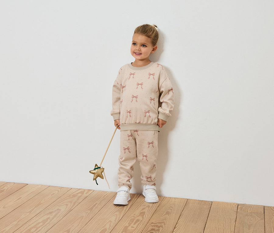 Usmívající se holčička drží hůlku ve tvaru hvězdy a má na sobě béžovou soupravu se vzorem mašlí.