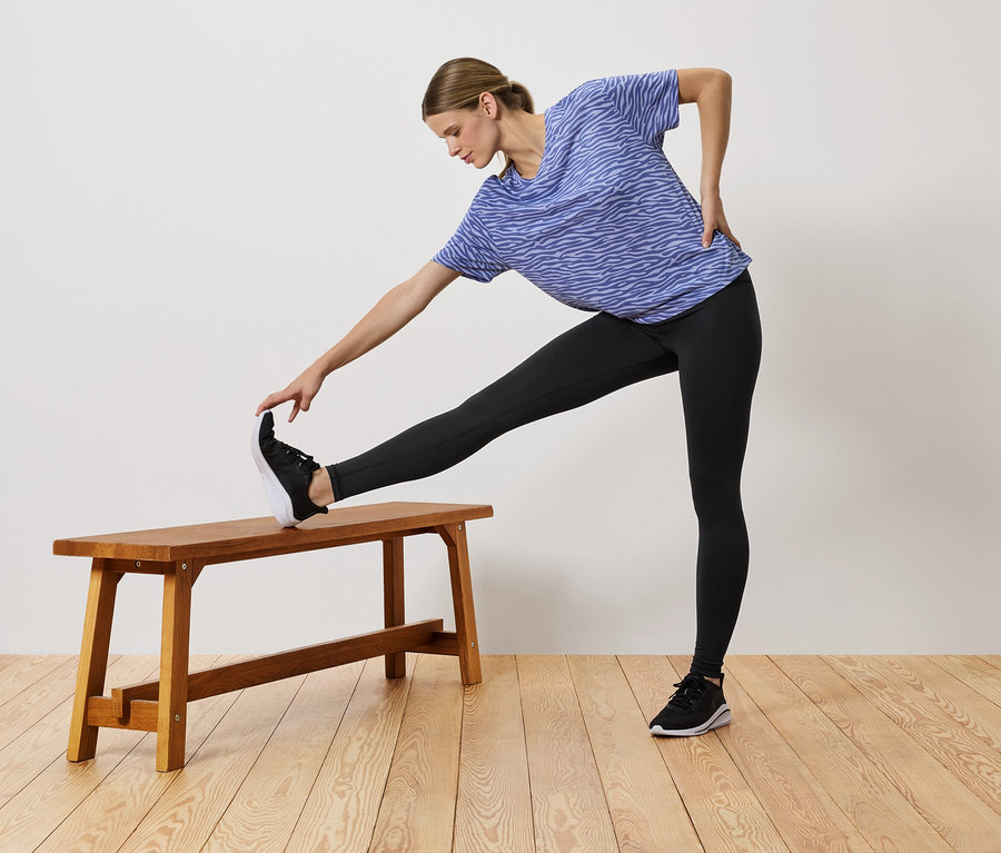 Žena v modrém tričku a černých legínách se protahuje s nohou na dřevěné lavici.