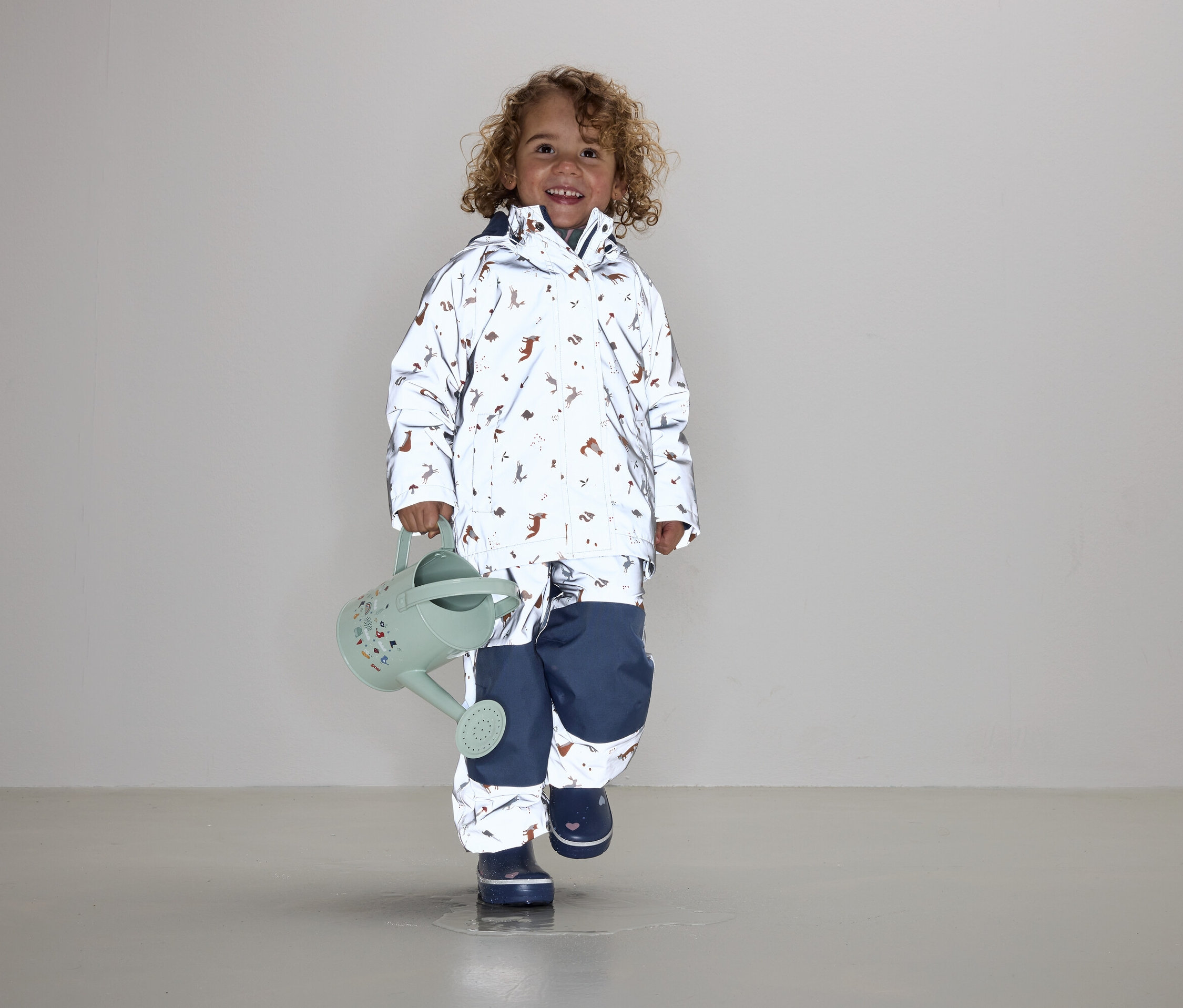 Dítě s kudrnatými vlasy v bundě a kalhotách s potiskem zvířat drží zelenou konev.