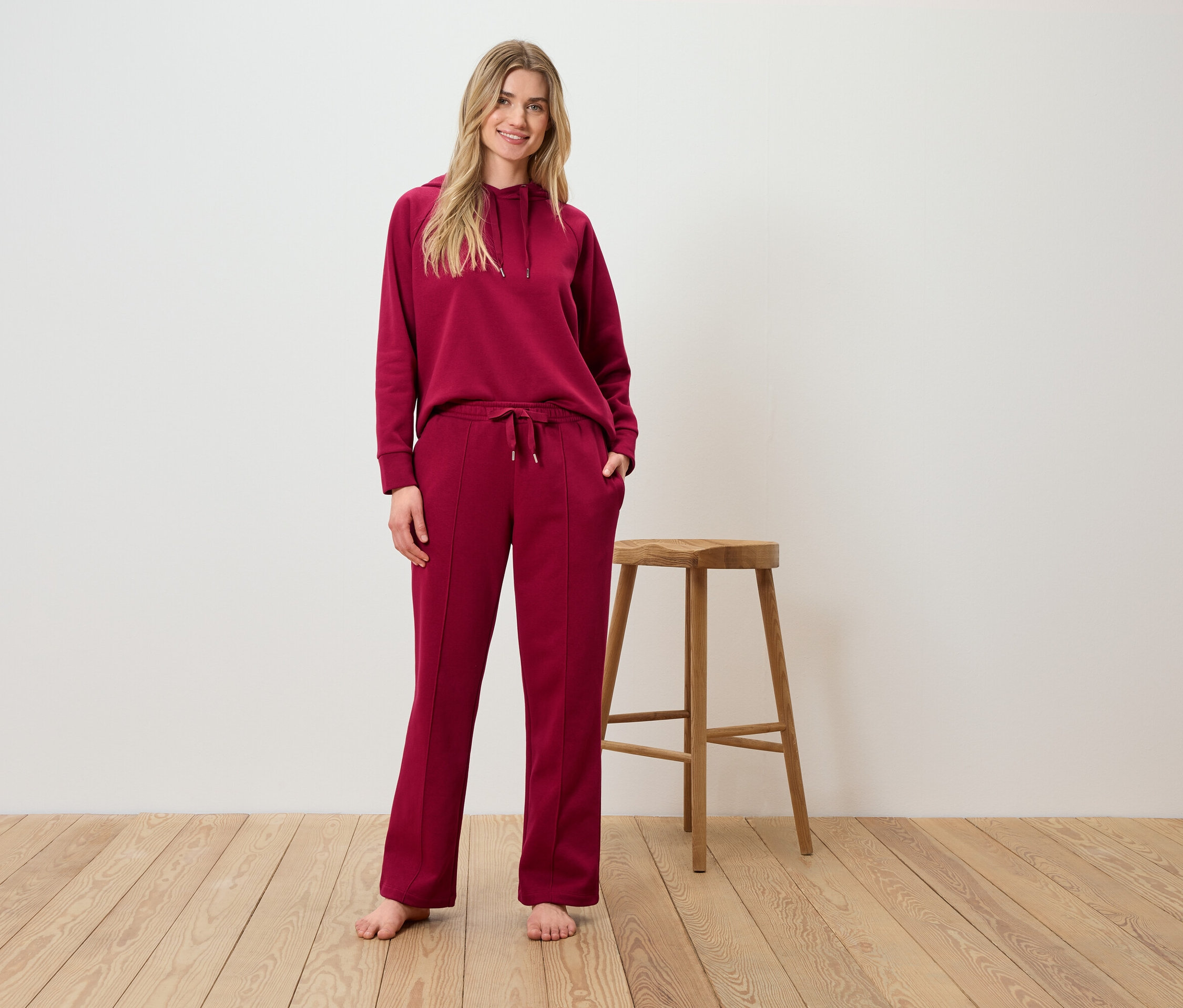 Žena pózuje v bordó soupravě s kapucí a kalhotami před dřevěnou stoličkou.