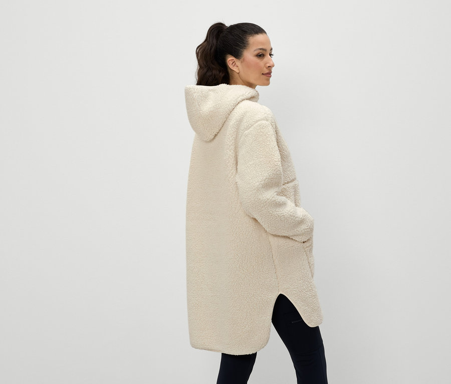 Žena z pohledu zezadu má na sobě krémový kabát s kapucí a ruce v kapsách.