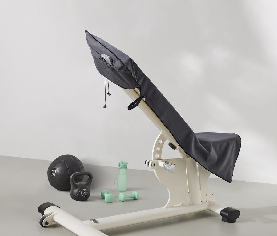 Posilovací lavice je zakrytá šedou látkou. Vedle lavice leží medicinbal, kettlebell, láhev na vodu a činky.