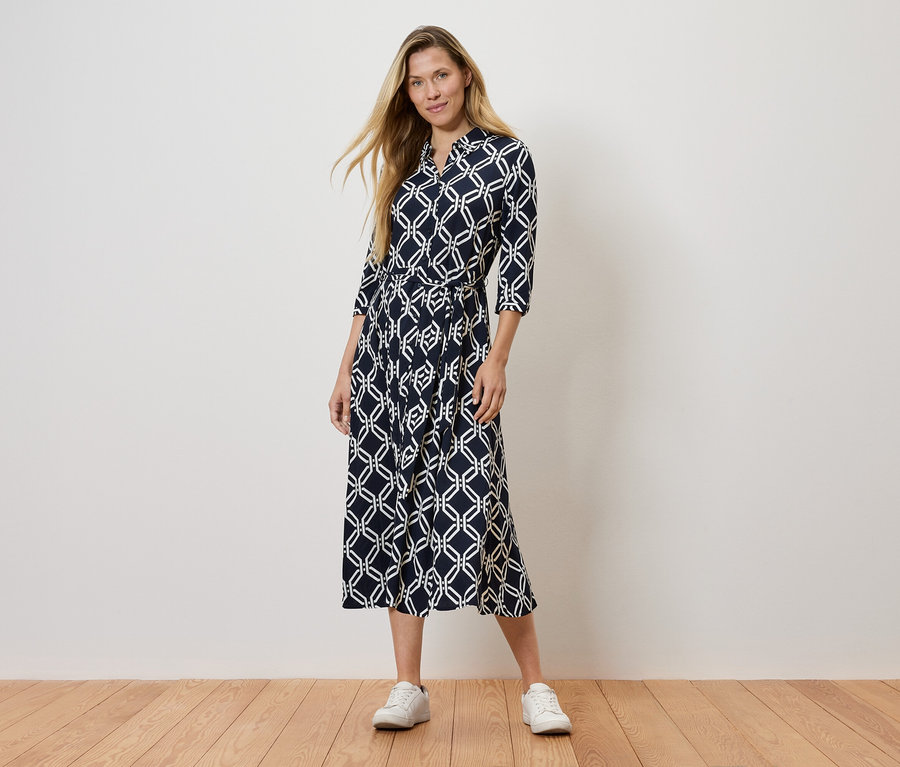 Žena stojí a má na sobě tmavě modré šaty se vzorem a bílé tenisky.
