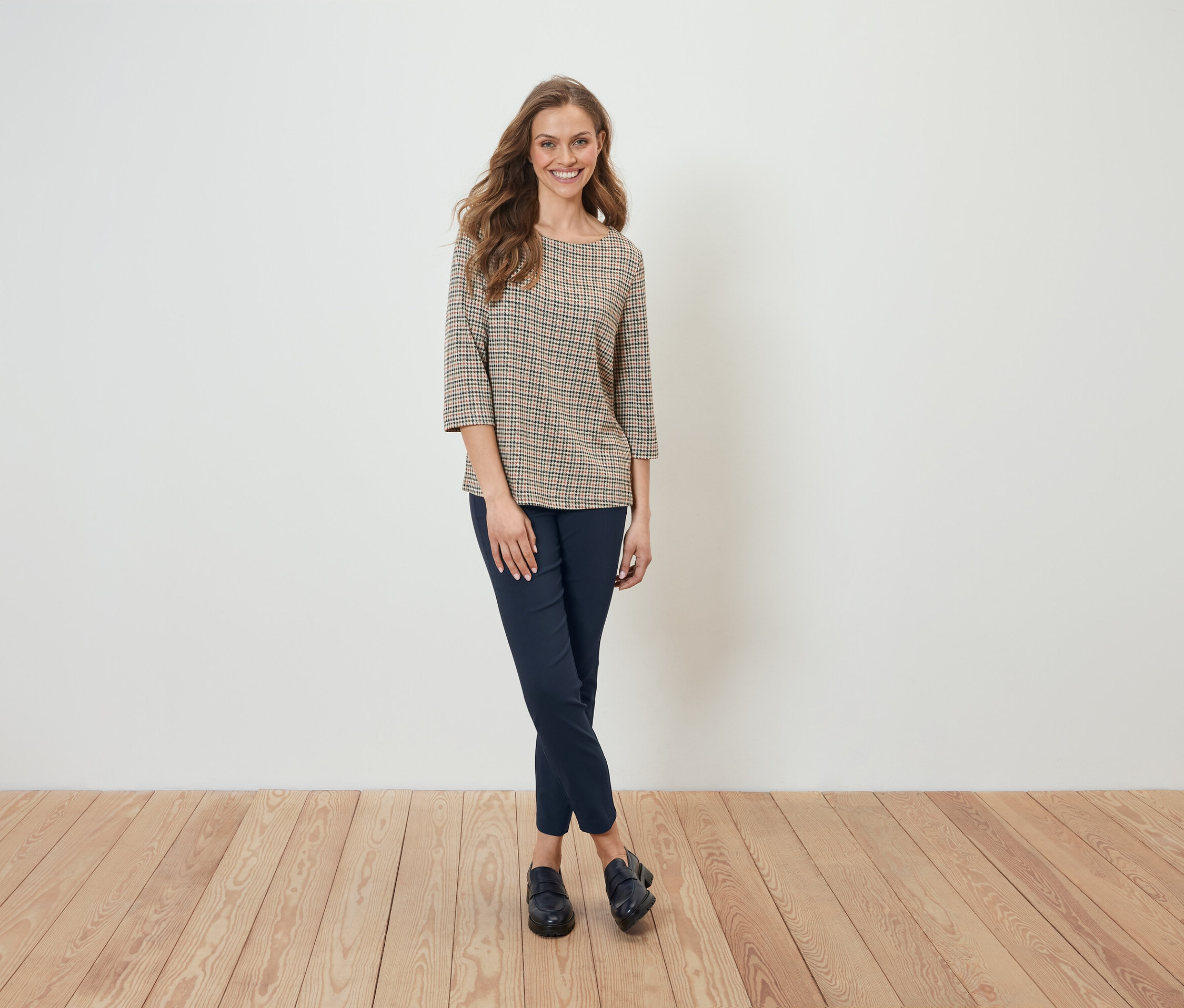 Žena v kostkované halence a tmavě modrých kalhotech stojí před bílou stěnou.