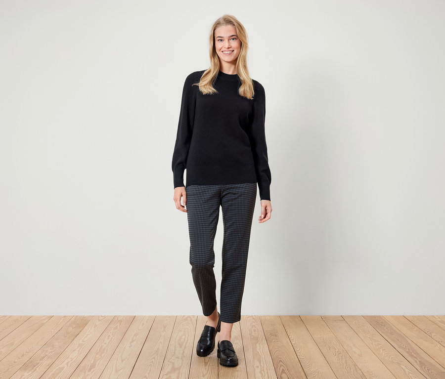 Žena stojí a usmívá se, má na sobě černý svetr, vzorované kalhoty a černé mokasíny.