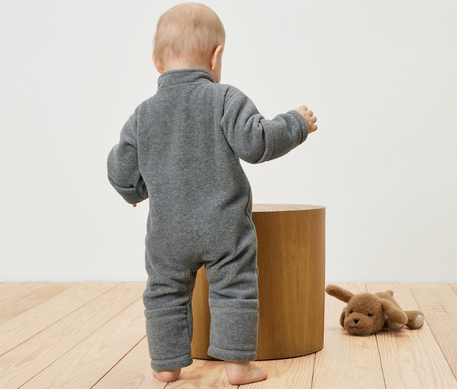 Batole v šedém fleecovém overalu stojí u dřevěné stoličky. Vedle leží plyšový pes.