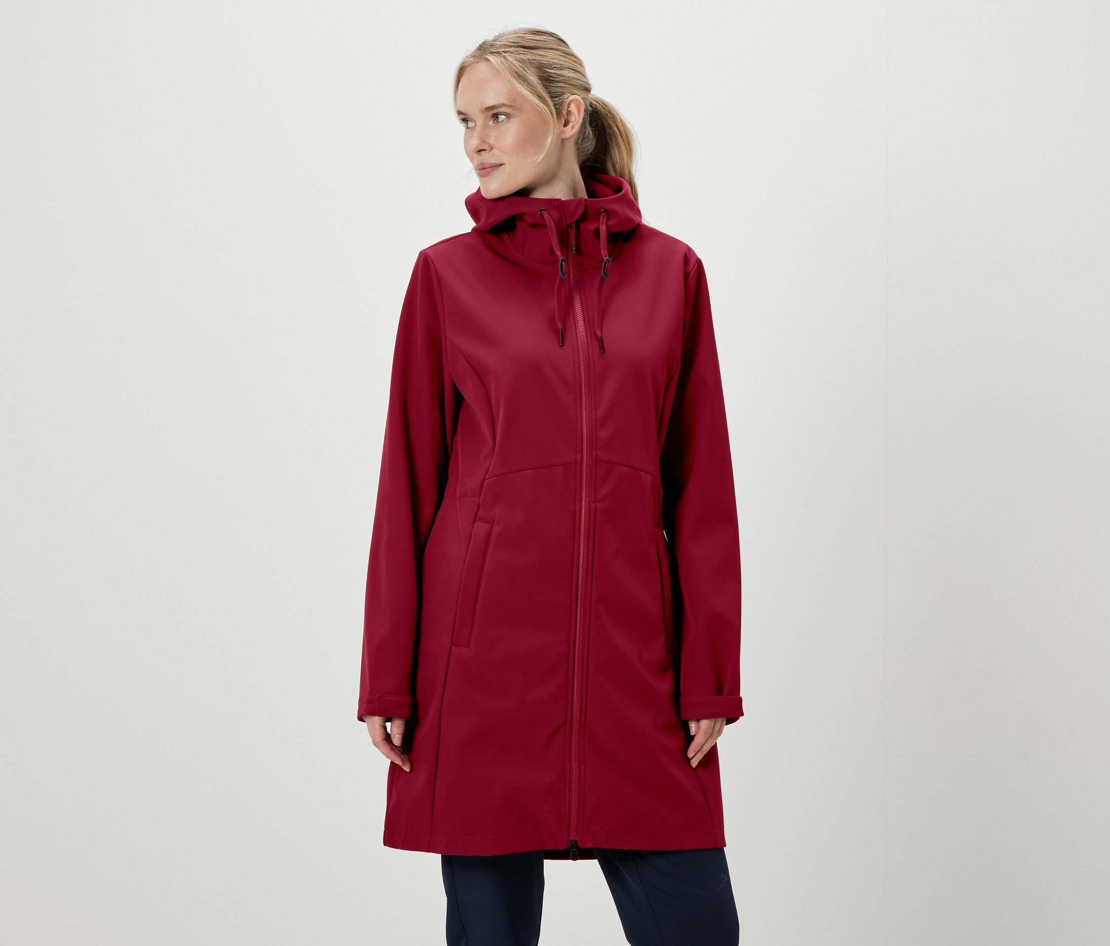 Blondýna v bordó bundě s kapucí na zip a kalhotách na bílém pozadí.