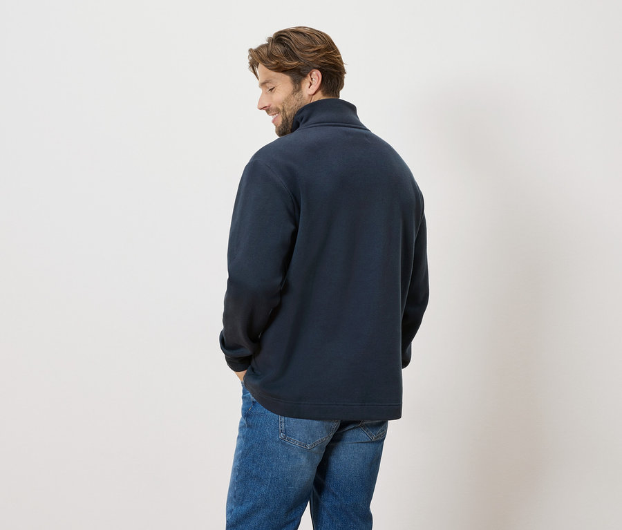 Muž stojí zády ke kameře, má na sobě tmavě modrý svetr a džíny.